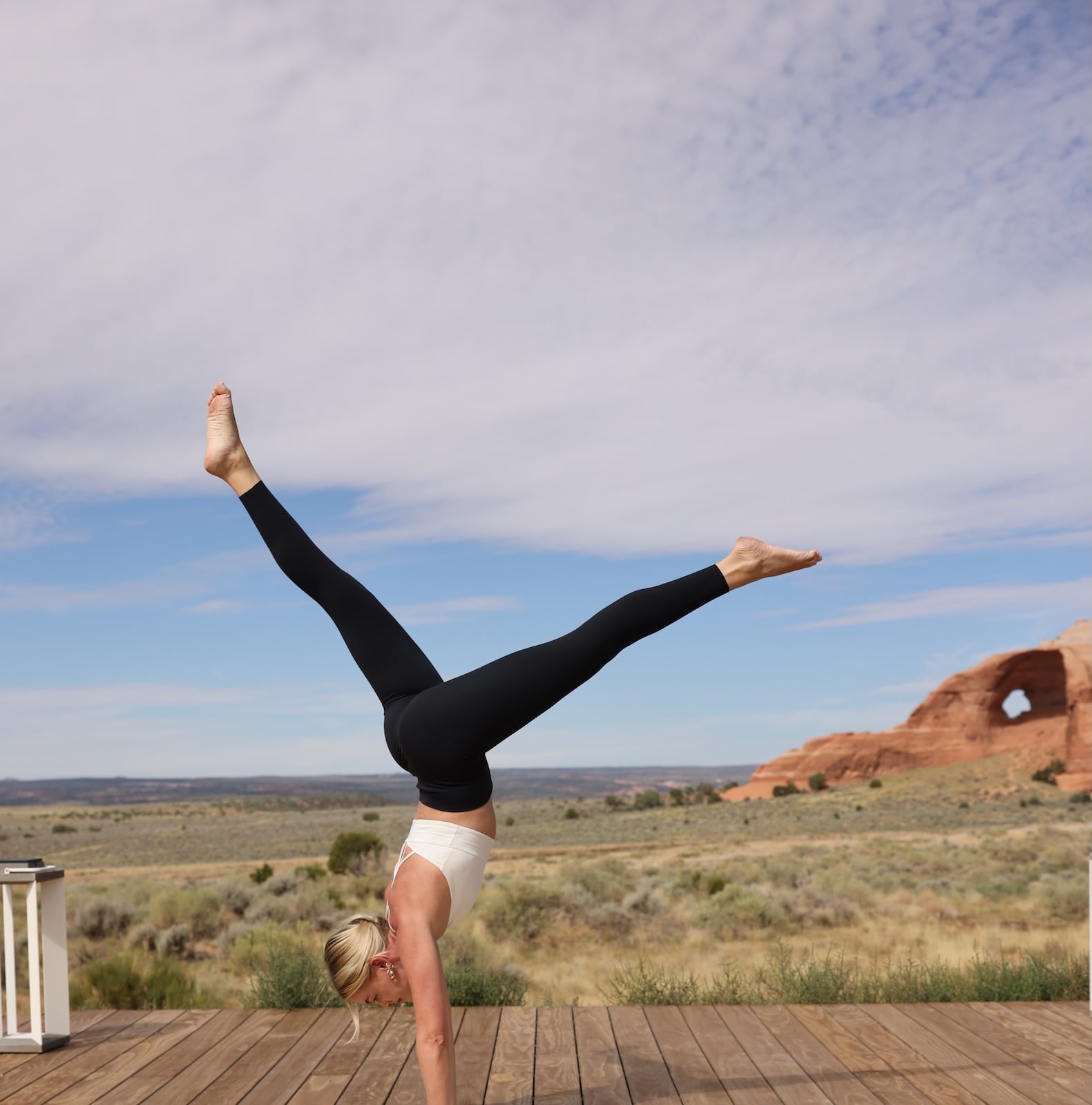 A woman in a handstand position on a wooden deck overlooking a desert landscape with a notable arch in the distance.