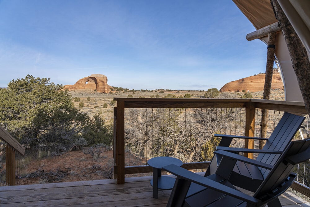 A balcony with two chairs and a table overlooking a desert landscape with a large rock formation.