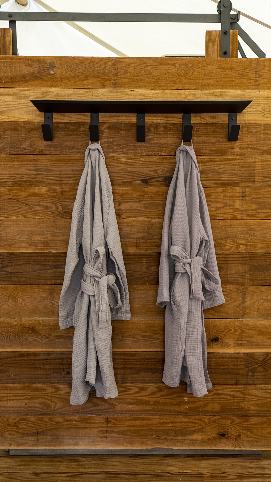 Two gray bathrobes hang from hooks on a wooden wall.