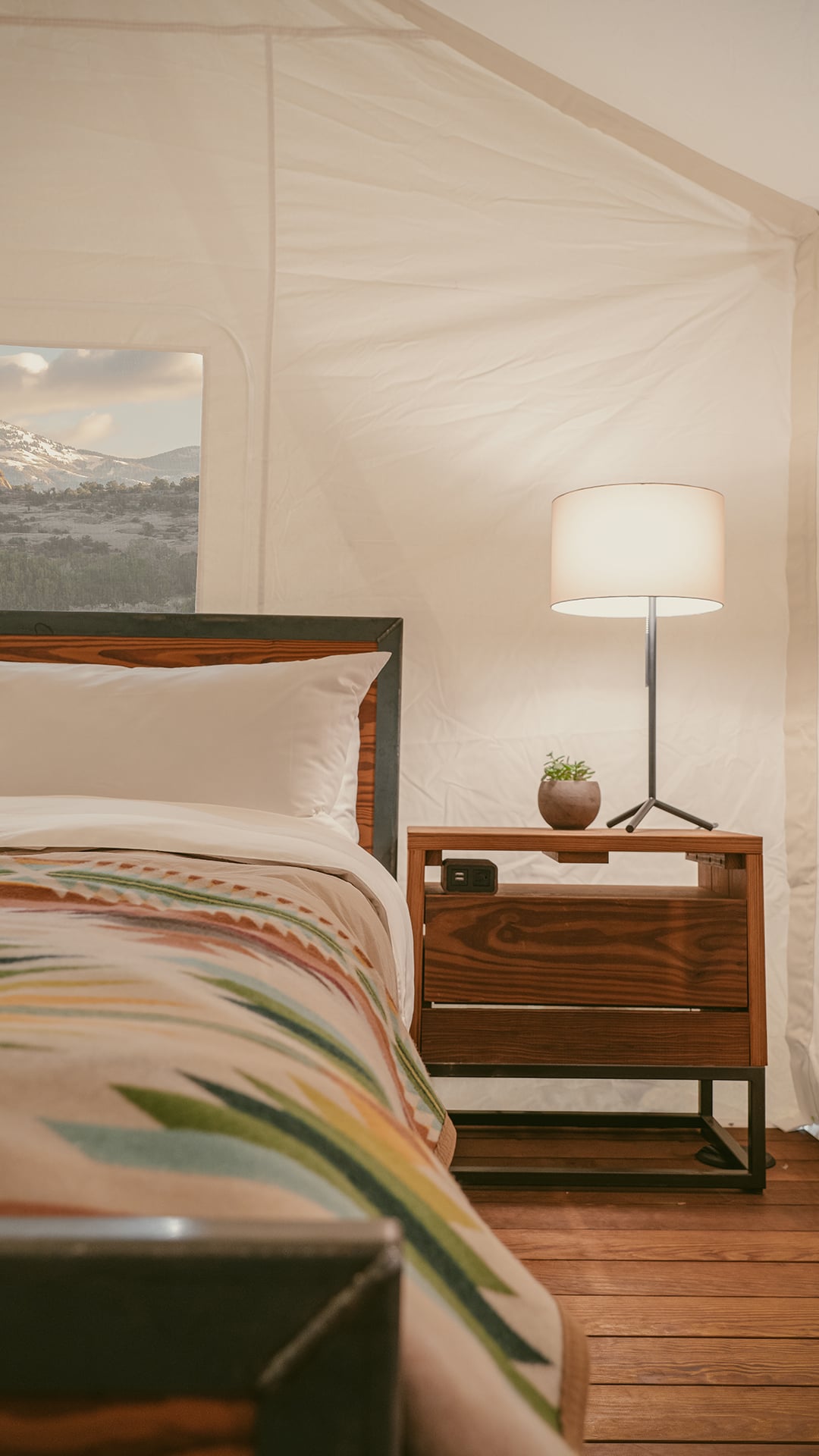 A close-up view of a bed with a colorful blanket, white pillows, and a wooden nightstand with a lamp and small potted plant.
